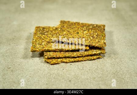 Vue latérale rapprochée d'une pile de craquelins de graines artisanales ou de pain croustillant placés sur une surface texturée beige neutre. Les craquelins rectangulaires présentent un Banque D'Images