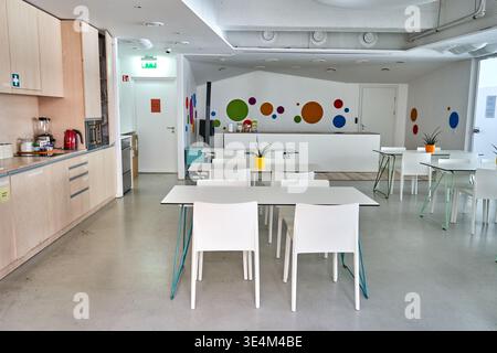 Intérieur moderne et lumineux de cafétéria ou de salle de pause avec tables et chaises blanches, armoires de cuisine en bois clair et mur circulaire coloré Banque D'Images