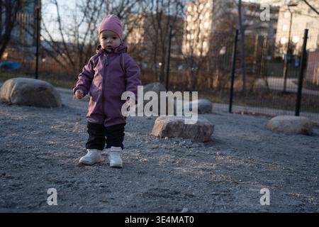 enfant en bonnet rose et combinaison de neige violette debout sur une aire de jeux de gravier au crépuscule près de grands rochers et clôture métallique, lumière du soleil dorée douce sur apa urbain Banque D'Images
