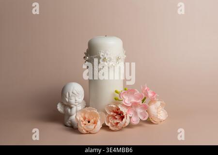 Une bougie décorative blanche avec des détails de dentelle et de perles est placée à côté d'une petite figurine d'ange et plusieurs fleurs artificielles rose clair sur un BA beige Banque D'Images