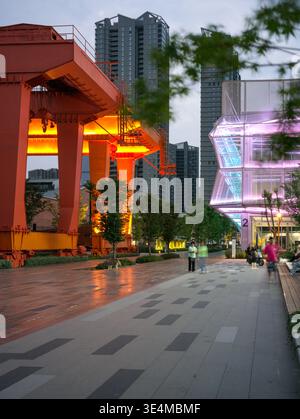 Wuhan, Chine - 11 juin 2025 : grande structure de grue industrielle orange illuminée de nuit au-dessus d'une passerelle piétonne pavée dans un parc urbain. Banque D'Images