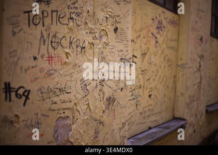 Gros plan en perspective d'un mur vieilli texturé avec de la peinture jaune écaillée. La surface est fortement recouverte de graffitis divers, y compris des noms Banque D'Images
