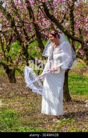 Belle mariée portant une robe blanche, voile, et couronne de fleur, tenant un parasol de dentelle délicate, célébrant la romance et l'amour dans une pêche rose vibrante b Banque D'Images