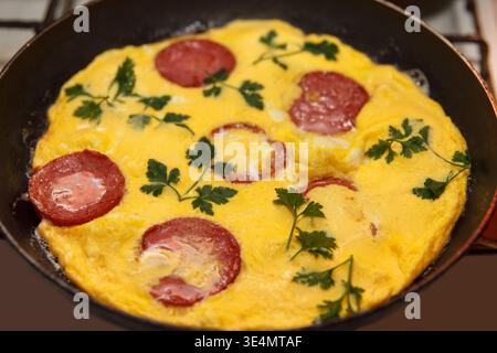 Une omelette dorée cuite dans une poêle, garnie de tranches de saucisse et de persil frais. Le plat est placé sur une cuisinière Banque D'Images