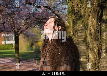 Jeune femme debout sous un arbre en fleurs de cerisier profitant de la lumière chaude du soleil avec les yeux fermés. Style de vie printanier et humeur paisible. Banque D'Images