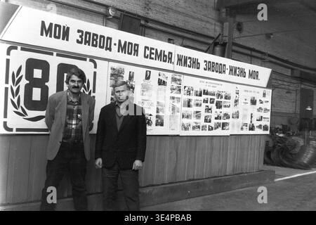 Sloviansk, 1995. Installation de raccords et isolateurs (AIZ). Deux employés masculins se tiennent debout dans la zone du comité syndical à côté d'un grand tableau d'affichage d'entreprise avec la mention « My Plant - My Family ». Le stand visuel présente des photographies d'archives célébrant le quatre-vingtième anniversaire de l'entreprise. Cette photographie inspirante du milieu des années 90 capture magnifiquement la forte identité industrielle, la fierté collective et le profond respect pour le patrimoine de travail, documentant la vie communautaire dynamique et la culture du Donbass paisible. Banque D'Images