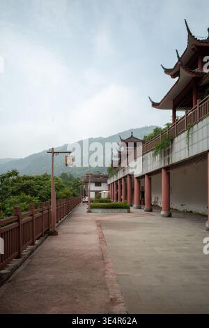 L'architecture chinoise ancienne borde un chemin de cour tranquille. Les piliers rouges soutiennent les gouttières inclinées traditionnelles et les garde-corps en bois. Collines verdoyantes s'élèvent Banque D'Images