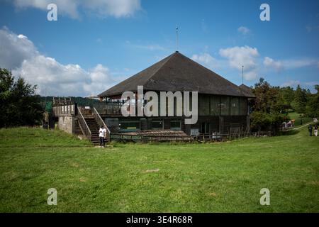 Lodge en bois avec toit de chaume se dresse sur une colline herbeuse. Le ciel bleu avec des nuages fournit une toile de fond lumineuse. Terrasse en bois et escaliers offrent un accès à la s. Banque D'Images