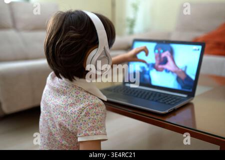 Geste joyeux de jeunesse. Fille heureuse portant des écouteurs faisant des signes de coeur à son ordinateur portable dans une chambre confortable Banque D'Images