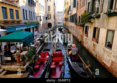 Venise, Italie-28e septembre 2025 : gondoles dans l'étroit canal vénitien, gondolier rame près des bâtiments historiques et un restaurant en plein air au bord de l'eau. Banque D'Images