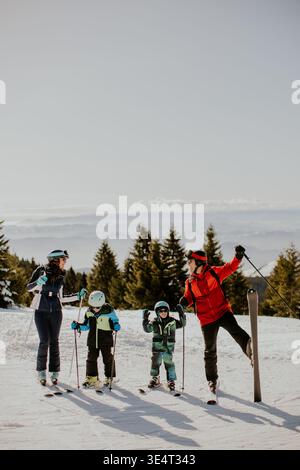 Une famille de quatre personnes se tient sur une pente enneigée profitant du ski. Deux enfants tiennent des bâtons de ski tout en souriant. Les adultes les guident. Arbres couverts de neige surroun Banque D'Images