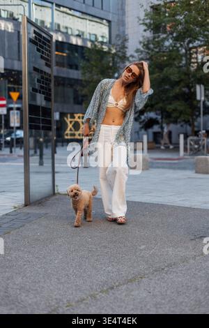 Jeune femme promenant son petit chien bouclé dans un cadre urbain moderne. Elle porte des vêtements d'été décontractés et des lunettes de soleil, profitant d'une promenade détendue. Urban Lif Banque D'Images