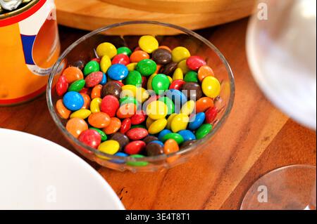 Un bol en verre transparent rempli de morceaux de chocolat recouverts de bonbons colorés sur une table en bois Banque D'Images
