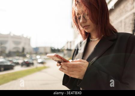 Jeune femme à la mode texto sur smartphone dans la rue de la ville. Style de vie moderne, communication numérique, fond urbain, tenue élégante. Banque D'Images