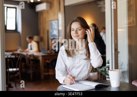 Étudiant ou freelance écrivant dans un cahier à la table du café. Cadre décontracté et moderne avec tasse à café et smartphone. Parfait pour l'éducation, à distance Banque D'Images