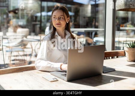 Jeune femme confiante travaillant sur un ordinateur portable dans un café moderne et lumineux. Portant une chemise blanche, elle est assise à une table en bois avec de la lumière naturelle et su élégant Banque D'Images