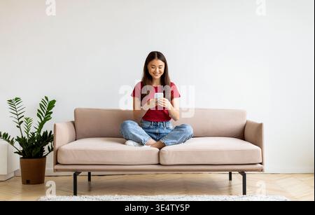 Souriant jeune femme asiatique avec smartphone se relaxant sur Couch à la maison Banque D'Images