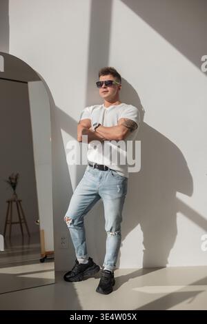 Portrait d'un homme élégant en lunettes de soleil, posant décontractée à l'intérieur. Vêtu d'un tee-shirt blanc et d'un denim léger, avec un bras tatoué, il capture un mod audacieux Banque D'Images
