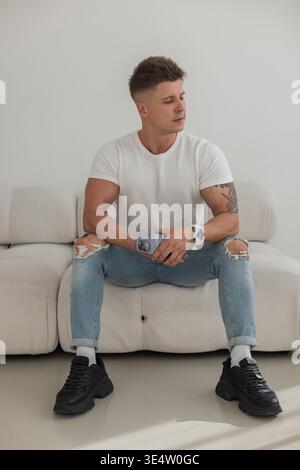 Élégant bel homme à succès avec coiffure en T-shirt blanc avec jeans déchiré de mode et baskets avec montre tient smartphone et se trouve sur le canapé en R. Banque D'Images