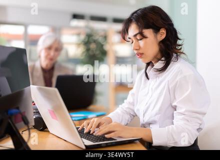 Jeune femme assise au bureau et tapant sur ordinateur portable dans un bureau moderne Banque D'Images