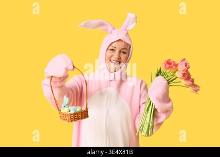 Femme senior drôle en costume de lapin tenant le panier avec des œufs de Pâques et des tulipes sur fond jaune Banque D'Images