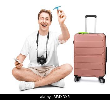 Heureux voyageur masculin assis avec une valise rose, tenant un avion en papier et un smartphone. Touriste excité avec appareil photo prêt pour le voyage de vacances. Isolat Banque D'Images
