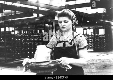 Sloviansk, 1950s-1960s, armature-Insulator Plant (AIZ). Une ouvrière d'usine portant un foulard traditionnel nettoie méticuleusement et inspecte un grand isolant électrique vitré brun foncé. La photographie d'archives en noir et blanc capture l'environnement de l'atelier d'assemblage, avec des systèmes de convoyeurs aériens et des racks de composants de lignes électriques robustes. Ce portrait industriel authentique reflète le travail manuel dévoué des femmes soviétiques, préservant l'histoire de la fabrication et la vie du Donbass pacifique. Banque D'Images