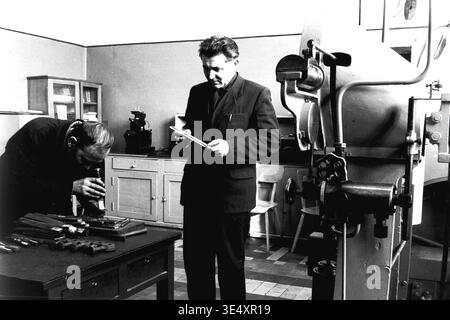 Sloviansk, 1950s-1960s, armature-Insulator Plant (AIZ). Deux spécialistes techniques effectuent un contrôle qualité rigoureux sur les composants en métaux lourds à l'intérieur d'un laboratoire d'usine lumineux. Un ingénieur se tient debout pour examiner une pièce, tandis que son collègue assis utilise un instrument grossissant pour inspecter un lot de matériel fabriqué sur le bureau en bois. Une grande machine d'essai industrielle se trouve à proximité. Cette scène historique met en valeur le travail méticuleux de l'ingénierie, préservant l'atmosphère authentique de la vie avant la guerre et le patrimoine industriel d'un Donbass paisible. Banque D'Images