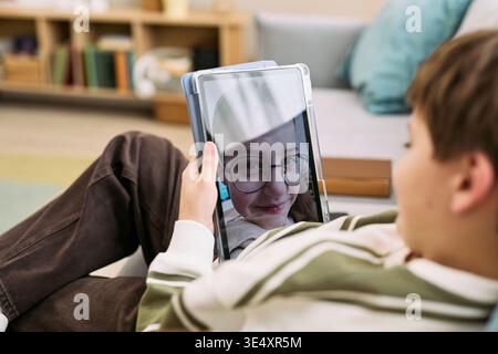 Garçon caucasien allongé sur un canapé tenant une tablette montrant un appel vidéo avec une fille caucasienne portant des lunettes souriant à l'écran, capturant la communication de l'enfance et l'interaction numérique à la maison Banque D'Images