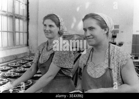 Sloviansk, 1950s-1960s, armature-Insulator Plant (AIZ). Deux ouvrières d'usine organisent des isolateurs électriques en céramique sombre fraîchement émaillés le long d'un séchoir mécanisé placé à côté d'une grande fenêtre d'atelier. Travaillant côte à côte dans leurs tabliers industriels, ils assurent un flux régulier et continu de composants lourds à travers le processus de fabrication exigeant. Cette photographie documentaire expressive capture de manière éclatante le travail authentique des femmes, honorant la mémoire de la vie quotidienne avant la guerre et un Donbass pacifique. Banque D'Images