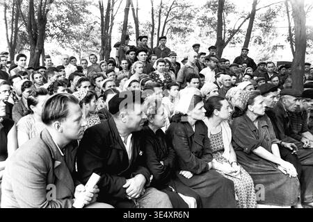 Sloviansk, 1950s-1960s, armature-Insulator Plant (AIZ). Une grande foule de travailleurs attentifs de l'usine se réunit à l'extérieur dans la cour industrielle ombragée pour une réunion collective du personnel. Les hommes et les femmes de la classe ouvrière sont assis étroitement ensemble sur des bancs en bois, écoutant attentivement un haut-parleur hors caméra. Cette photographie documentaire authentique capture le fort esprit communautaire des entreprises soviétiques, honorant la mémoire vitale de la vie quotidienne avant la guerre et l'héritage durable d'un Donbass pacifique. Banque D'Images
