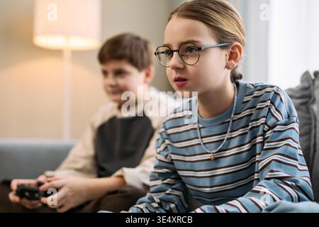 Caucasien preteen girl portant des lunettes assis sur le canapé jouant à un jeu vidéo avec le caucasien preteen boy en arrière-plan, tenant le contrôleur de jeu à la fois concentré sur l'écran de télévision Banque D'Images