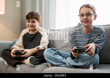 Deux enfants caucasiens assis sur le canapé jouant au jeu vidéo avec des contrôleurs, garçon et fille se concentrant sur l'écran, à la fois engagés et souriants, environnement familial décontracté, posture détendue Banque D'Images