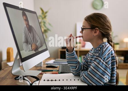 Fille caucasienne assise au bureau regardant la leçon en ligne sur le moniteur d'ordinateur, tenant le stylo près de la bouche, se concentrant sur l'enseignant masculin à l'écran, entouré de cahiers et de matériel d'étude Banque D'Images