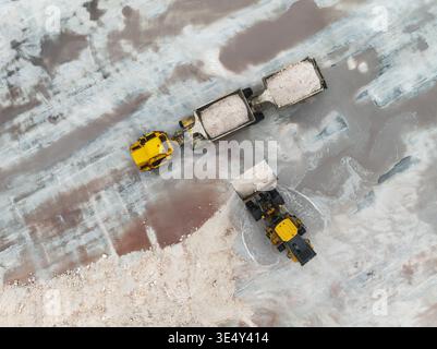 Vue aérienne de machines jaunes contrastant avec des marais salés pâles, un ballet industriel brutal se déroule, Walvis Bay, région d'Erongo, Namibie. Banque D'Images