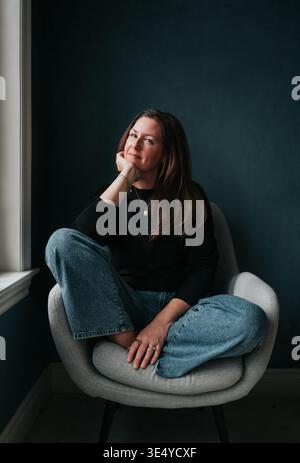 Belle femme d'âge moyen assise dans une chaise à côté de la fenêtre. Banque D'Images