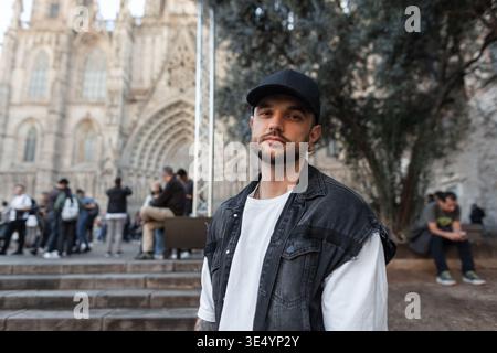 Bel homme hipster de mode avec casquette noire avec t-shirt blanc avec gilet en denim marchant à Barcelone près de l'église Banque D'Images