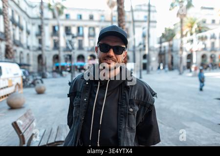 Cool bel homme à la mode dans des vêtements en denim noir avec veste avec casquette avec lunettes de soleil marchant à Barcelone dans la ville Banque D'Images