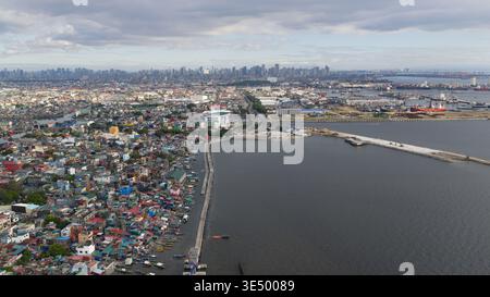 Vue aérienne de logements côtiers denses et de la zone portuaire animée avec l'horizon lointain de la ville sous un ciel nuageux à Navotas, Metro Manila, Philippines. Banque D'Images