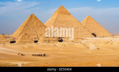 Vue complète des trois principales pyramides de Gizeh (Khufu, Khafre, Menkaure) avec caravanes de chameaux traversant le désert en face Banque D'Images