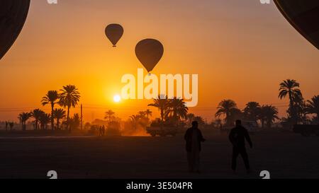 Deux montgolfières silhouettées contre un soleil orange vif, des palmiers et des populations locales au premier plan, Louxor, Egypte Banque D'Images