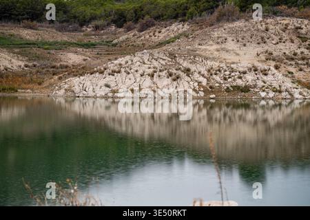 Une banque rocheuse avec des buissons clairsemés se reflète parfaitement dans l'eau encore verte d'un réservoir. L'image montre des motifs d'acros de pierre et de végétation Banque D'Images