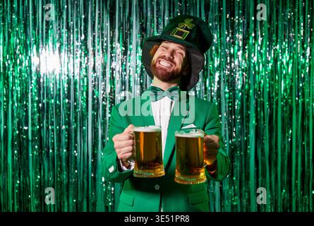 Homme heureux portant costume vert et chapeau tenant deux tasses de bière, posant contre vert brillant Banque D'Images