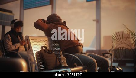 Voyageur afro-américain détendu s'étirant et regardant les avions par la fenêtre de l'aéroport. L'homme lève les bras pour étirer les muscles, soulager la tension du voyage, regarde par la fenêtre les avions sur la piste. Banque D'Images