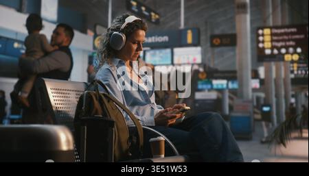 La femme de voyage élégante apprécie les listes de lecture préférées et parcourt le contenu numérique sur smartphone dans le salon Airport, en portant des écouteurs audio. Concept de la culture de voyage des jeunes et Tech Savvy Lifestyle. Banque D'Images
