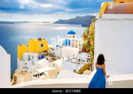 Une jeune femme en robe bleue se tient sur un escalier surplombant les églises au dôme bleu du village blanc d'Oia, sur l'île de Santorin, en Grèce Banque D'Images