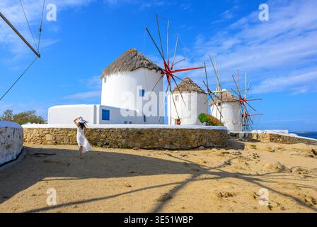 Une jeune femme tient son chapeau dans la brise des moulins à vent côtiers emblématiques de la ville de Mykonos, sur l'île de Mykonos, en Grèce. Banque D'Images