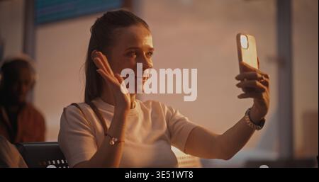 Assise avec la valise près, la femme s'engage dans un appel vidéo pendant l'escale. Femme regarde dans la caméra du téléphone, riant et discutant. Mode de vie des voyageurs connectés. Communications mobiles, connectivité 5G. Banque D'Images
