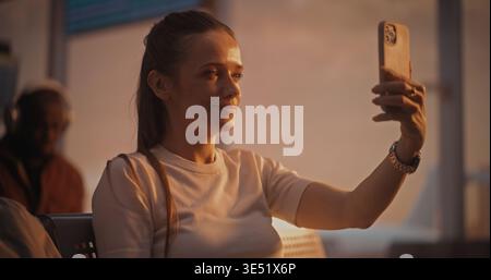 Assise avec la valise près, la femme s'engage dans un appel vidéo pendant l'escale. Femme regarde dans la caméra du téléphone, riant et discutant. Mode de vie des voyageurs connectés. Communications mobiles, connectivité 5G. Banque D'Images