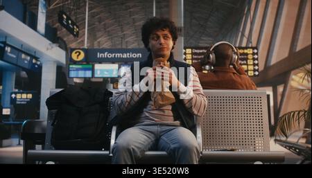 Jeune homme en chemise rayée et veste marine est assis dans le terminal de l'aéroport, dégustant la pâtisserie du sac en papier. Avec sac à dos sur le siège voisin, homme regarde autour lentement tout en mangeant. Moment de voyage fiable. Banque D'Images